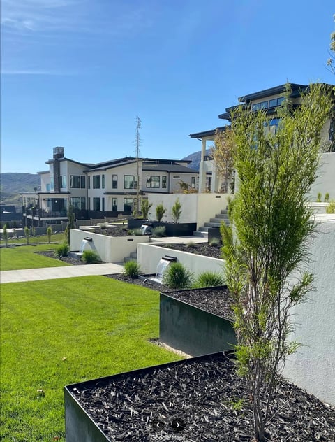 Modern residential complex with contemporary architecture, landscaped grounds featuring black planter boxes, green grass, and tall trees against a clear blue sky.