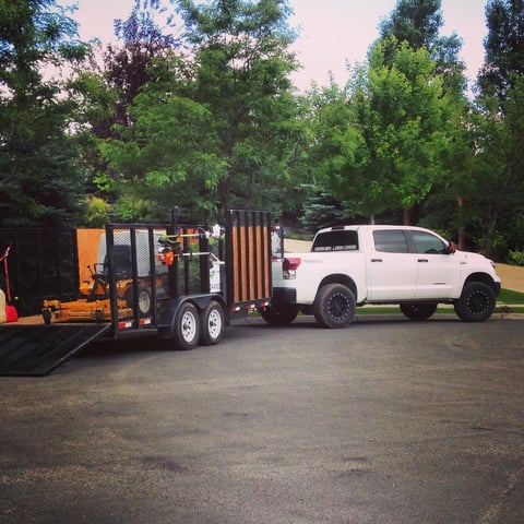 White pickup truck towing a black and orange enclosed equipment trailer in a park setting with green trees