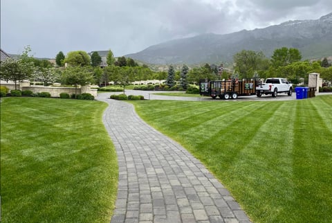Manicured lawn with gray stone pathway, work truck and equipment parked on right side, mountains visible in background
