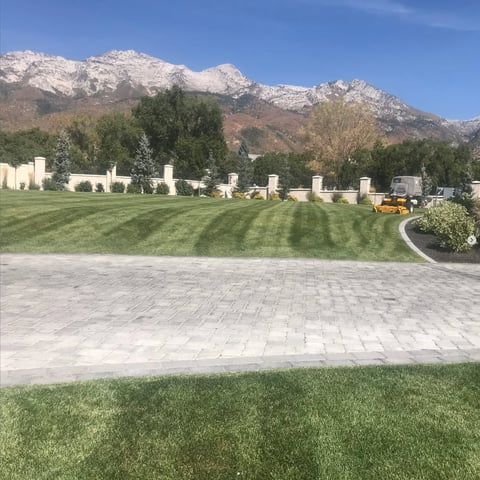 Cemetery with rows of headstones on green lawn, snow-capped mountains in background, and paved walkway in foreground