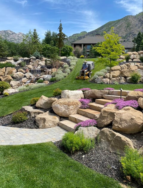 Landscaped backyard with large boulders, stone steps, purple flowers, manicured grass, and mountains in the background