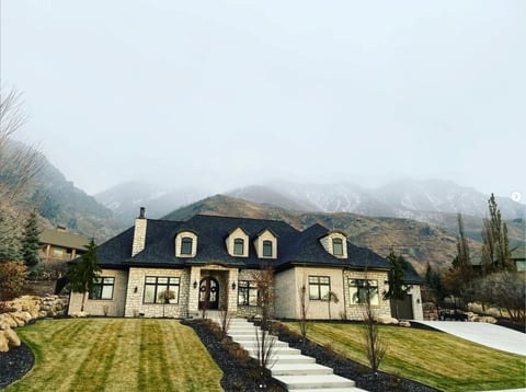Large two-story mansion with black roof and stone facade, manicured lawn, driveway, and snow-capped mountains in misty background