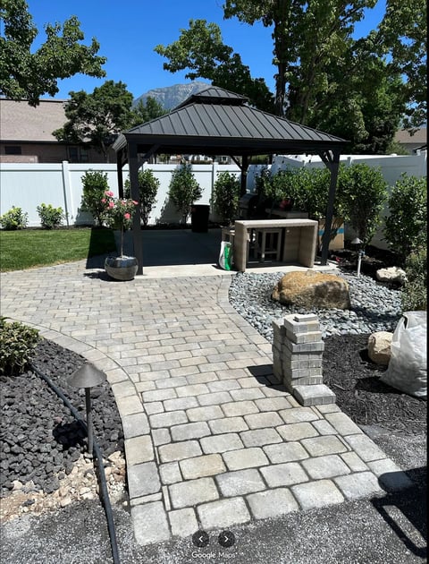 Modern backyard patio with gray metal gazebo, paver pathway, decorative rocks, and white privacy fence on sunny day