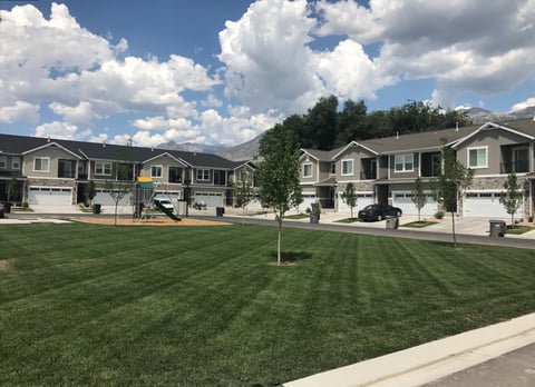 Row of newly constructed residential townhomes with garages lining a manicured green lawn under partly cloudy blue sky