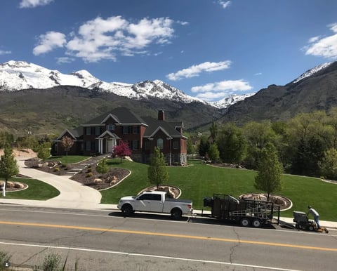 Large brick home on manicured lawn with snow-capped mountains in background, white truck and trailer parked on street